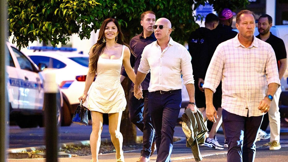 Lauren Sanchez wearing a yellow dress and Jeff Bezos wearing a white button down and dark pants while walking outside holding hands