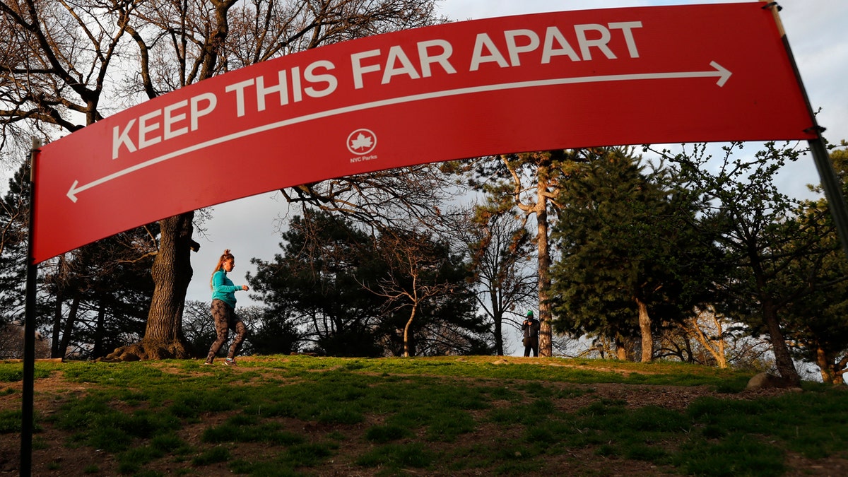 A woman jogs in Brooklyn's Fort Greeene park, Sunday, April 5, 2020, beneath a sign demonstrating the distance people should keep from each other during the coronavirus outbreak in New York.